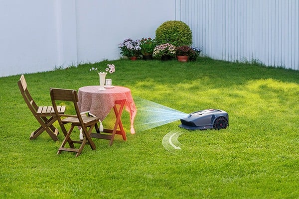 Na fotografii je robot, který seká trávu na zahradě. Zařízení se vyhýbá stolu se židlemi vedle něj.